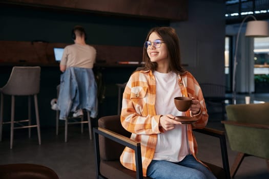 Pretty teenage girl resting on chair, drinking coffee. Attractive young lady worker chilling after work