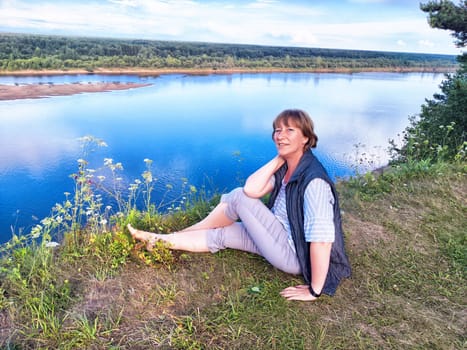 A middle-aged woman takes a selfie while relaxing by a tranquil river, surrounded by lush nature on a sunny day.