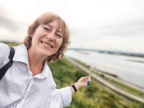 A middle-aged woman takes a joyful selfie with a river view, showcasing her love for travel and adventure.