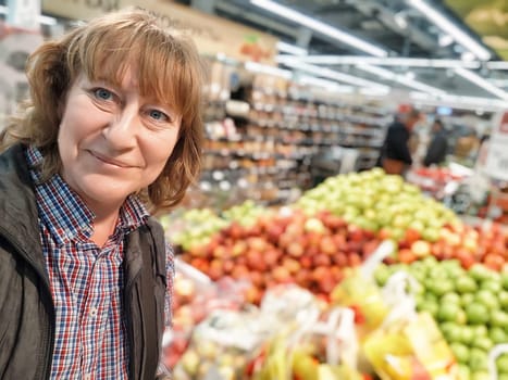A middle-aged female blogger smiles brightly while capturing a selfie surrounded by vibrant fruits in a grocery store.