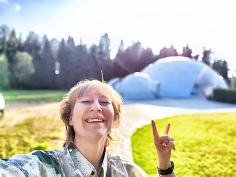 A middle-aged blonde woman smiles while taking a selfie near unique glamping accommodations in nature.