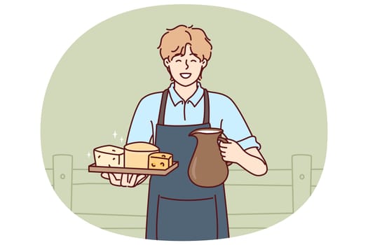 Man holds jug of milk and tray of cheese from cow farm selling own organic food. Guy wearing apron working on cow farm offers to buy organic dairy products with useful nutritional vitamins.