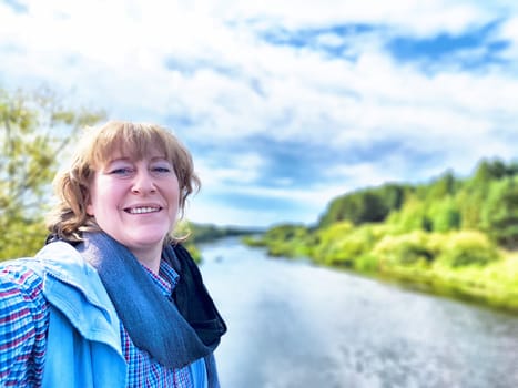A middle-aged traveler blogger enjoys her excursion by the river, capturing a cheerful selfie amidst nature's beauty.