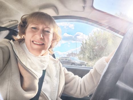 A middle-aged blonde woman smiles while driving, enjoying her adventure on a bright, sunny day.