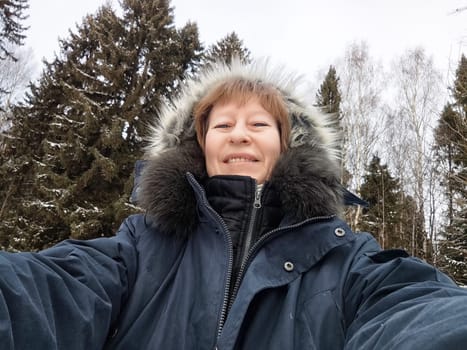Blonde woman smiles while taking a selfie in a snowy forest, showcasing her vacation spirit and love for adventure.
