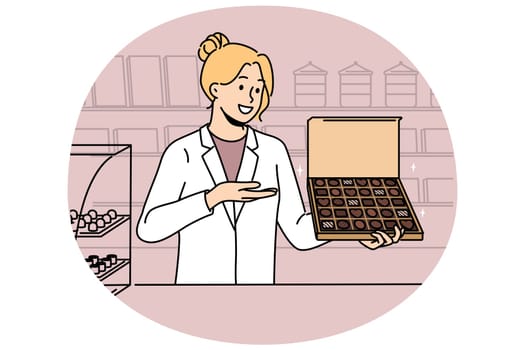 Woman seller with box of chocolates works in confectionery store and offers to buy appetizing dessert made from milk and cocoa. Girl in white coat of seller demonstrates chocolates with smile