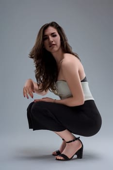Confident young woman crouching on grey background, exuding elegance in white top, black pants, and high heels, showcasing unique style and empowerment