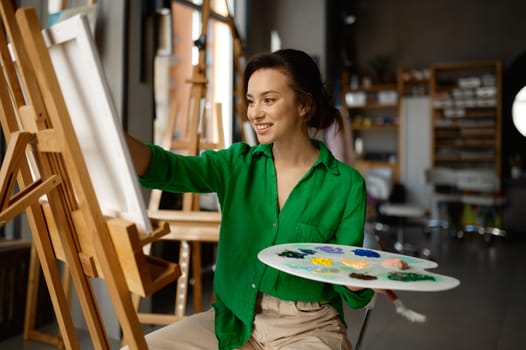 Portrait of happy young woman painting brush on canvas at workshop during art lesson class. Creative leisure activity concept