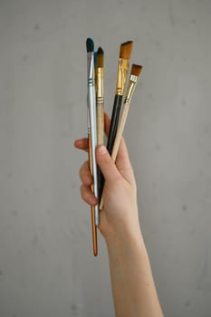 Female hand holding bunch of paintbrush over grey cement studio wall. Basic supplies for artist and art class school courses advertisement concept