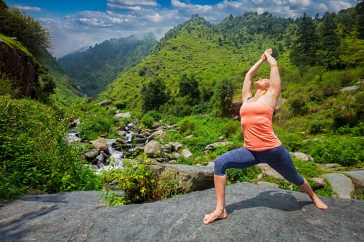 Yoga outdoors - woman doing Ashtanga Vinyasa Yoga asana Virabhadrasana 1 Warrior pose posture at waterfall in HImalayas mountains
