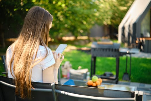 Rear side view of a woman relaxing on a bench with a smartphone in the backyard next to grill