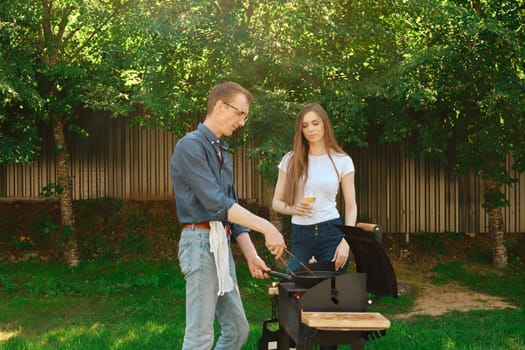 A middle-aged man cooks a beef steak on the grill for his daughter, who came to visit him at a country house
