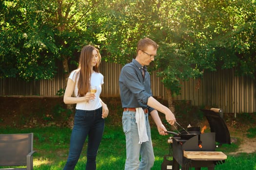 Middle-aged man grilling a beef steak on a frying pan on grill and his daughter watching on him standing at backyard