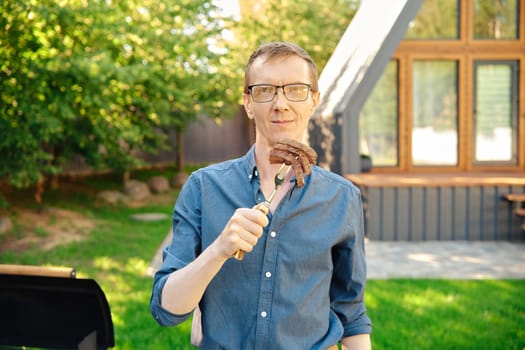 Middle-aged man standing at backyard of a summer house and holds a barbecue fork with slices of steak in his hand