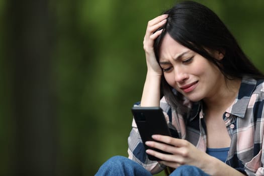 Worried asian woman checking bad news on cell phone and complaining in a park