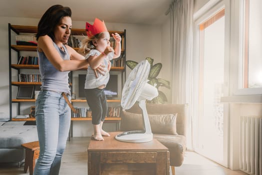 Mother lifts her daughter playing with a fan on a hot summer day at home, bonding and creating joyful memories in their cozy living room