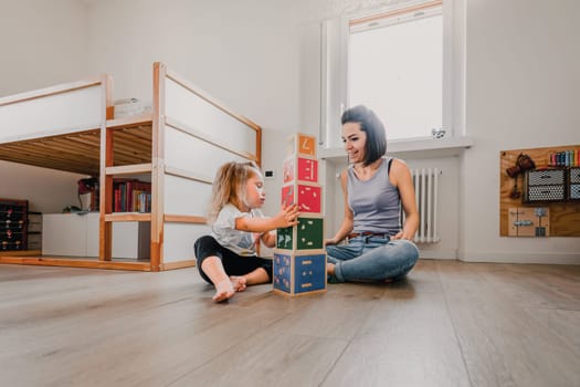 Mother and daughter sitting on a bedroom floor and playing together with building blocks, having fun, spending quality time together