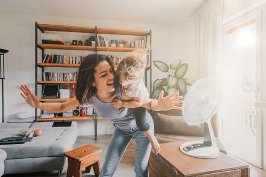 Mother and daughter beat the summer heat at home, playing with an electric fan, creating joyful memories in their cozy living room filled with warm natural light, embodying family togetherness