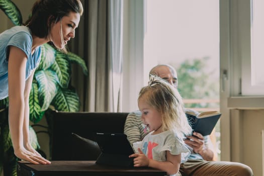 Young family is spending time together at home, with the mother watching over her daughter who is using a digital tablet while the father reads a book