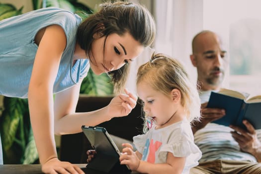 Young family is spending time together at home, with the mother showing something on a digital tablet to her daughter while the father is reading a book