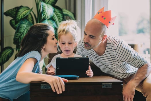 Young family is gathered around a digital tablet, enjoying their time together. The father is wearing a playful crown, adding to the cheerful atmosphere