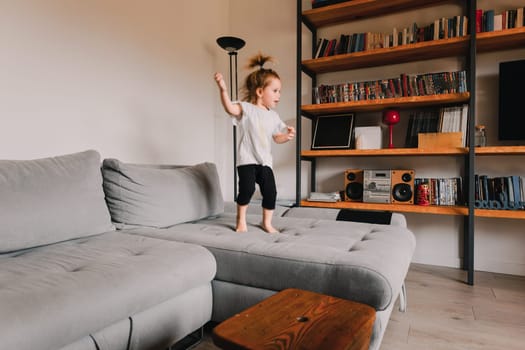 Happy girl jumps on a grey sofa in a bright living room, enjoying playtime at home