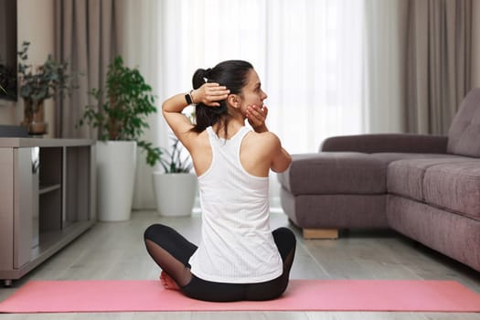 back view of Caucasian woman stretching head and warming up neck before workout sitting on yoga mat at home