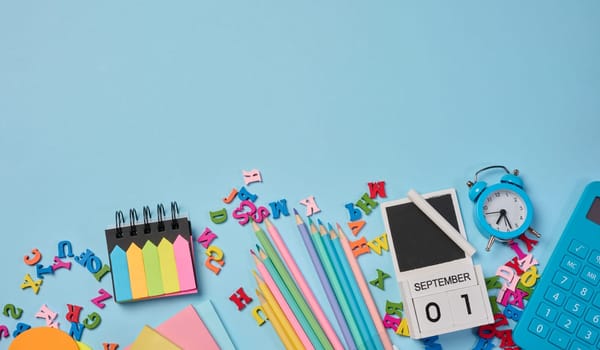Stationery laid out on a blue background. Multi-colored felt-tip pens, calculator, alarm clock, sticky notes and wooden letters, preparing for study or work, organizing workspace