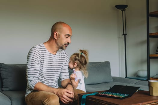 Young girl happily applies makeup to her father, playing together on the sofa with love and creativity. The cozy scene captures a special parent child bond in their home