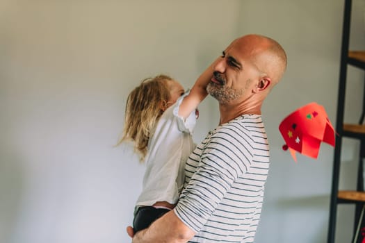 Young girl is holding a red hat and reaching up to touch her father's face as he holds her in his arms. They are both smiling and laughing, enjoying their time together