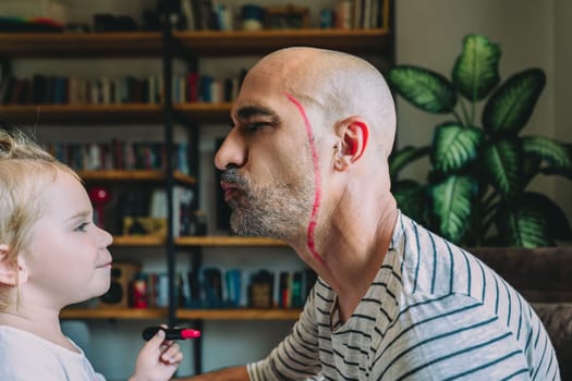 Girl playfully applying lipstick to her father's face, bonding in a cozy living room filled with books and plants. Celebrating family love and creativity