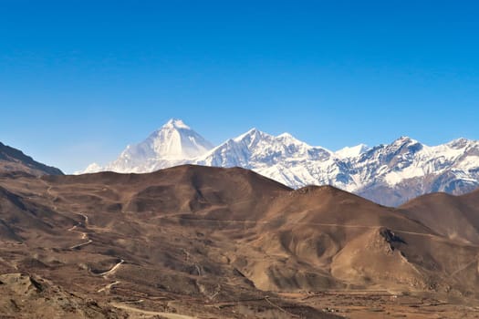 View onto the dry, hilly landscape that surrounds the town of Muktinath in front the snow covered Dhaulagiri I and Tukuche Peak, Annapurna Circuit Trek, Nepal 2022