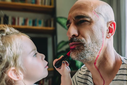 Little girl having fun putting lipstick on her father's face, who is patiently letting her do it, in a playful and loving atmosphere