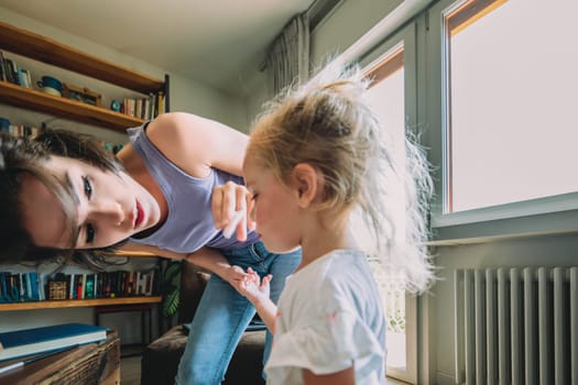 Mother is crouching and holding the hands of her little daughter, comforting her in their modern and bright living room