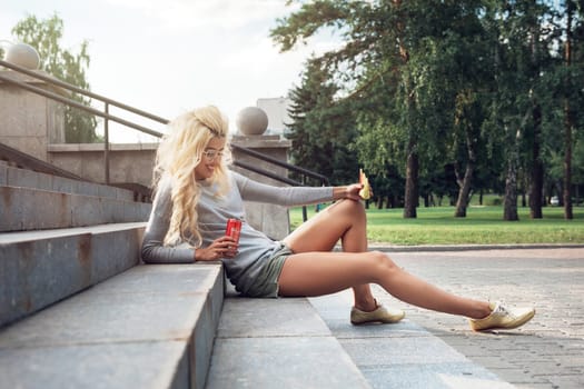 Rest in street, outdoor, people and leisure concept - beauty blonde young woman sitting on outdoor and eating