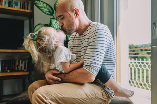 Bald man in a striped shirt and white pants kisses his little daughter on a bench surrounded by plants, books, and a window, showing their loving bond