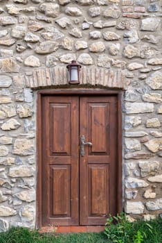 A wooden double door set within an arched stone wall with a lantern above. Green grass grows in front of the door