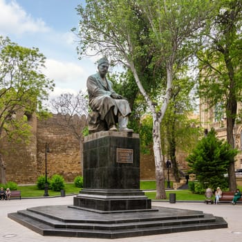 Baku, Azerbaijan - May 5, 2024: Bronze statue of Mirza Alakbar Sabir, a prominent Azerbaijani poet, sits in Sabir Garden. Behind the statue is the Old City stone wall, and lush greenery