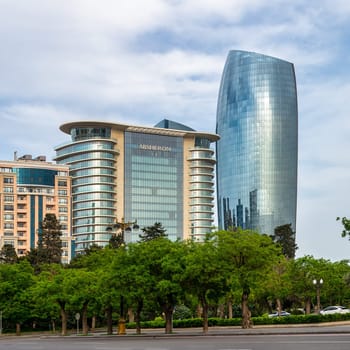 A view of modern highrise buildings in Baku, Azerbaijan, with a glassy, curved skyscraper in the background and a large building with green foliage in the foreground