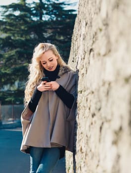 Stylish young tourist woman with long blonde hair using a smartphone app while leaning on an ancient stone wall in europe
