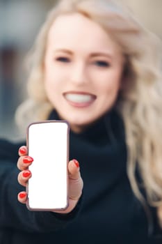 Cheerful woman proudly displays a smartphone with a clean white screen, perfect for showcasing your app or website. Beauty and fashion are emphasized in a casual, modern setting