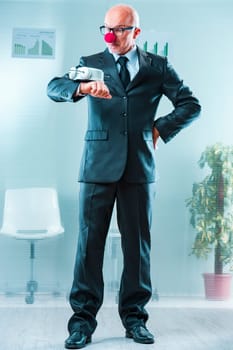 Gray-haired man in formal attire with a clown nose holds a large clock, symbolizing the ridiculousness of pressing deadlines due to poor management. Blurred office charts indicate a business environme
