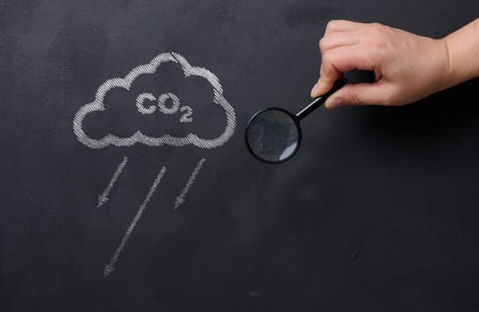 Female hand holds a magnifying glass against a black chalkboard background and a cloud, concept of reducing CO2 emissions