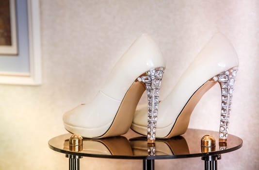 High-heeled wedding shoes decorated with transparent crystals stand on a glass transparent table