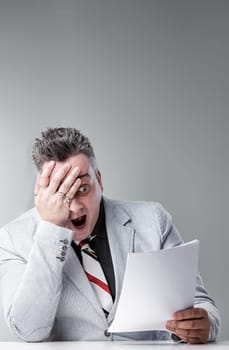 Businessman is sitting at a desk holding a document and looking shocked, with his hand over his face and mouth wide open