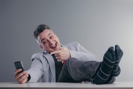 Businessman is sitting with his feet up on his desk, laughing at something on his phone. He is completely disregarding his work and acting unprofessionally