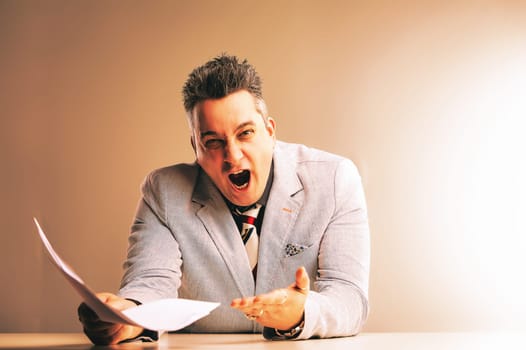 Businessman is experiencing extreme frustration, holding papers and shouting with his mouth wide open, conveying workplace stress and anger