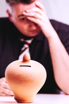 Businessman is having financial troubles as he sits at his desk with his head in his hand, an empty piggy bank in the foreground