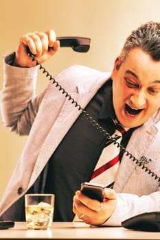 Businessman is holding a smartphone and screaming at a landline phone while drinking alcohol issues of alcoholism in the workplace