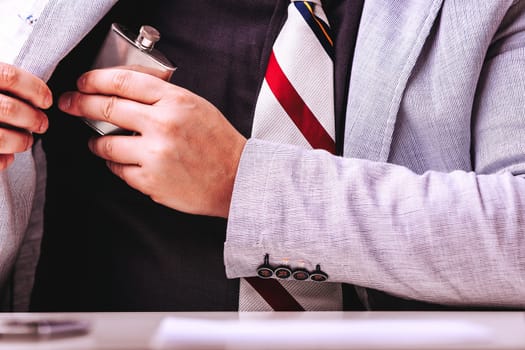 Businessman discreetly hides a flask in his suit pocket, coping with work stress through alcohol. His hand slips the bottle in, showing his unhealthy dependence